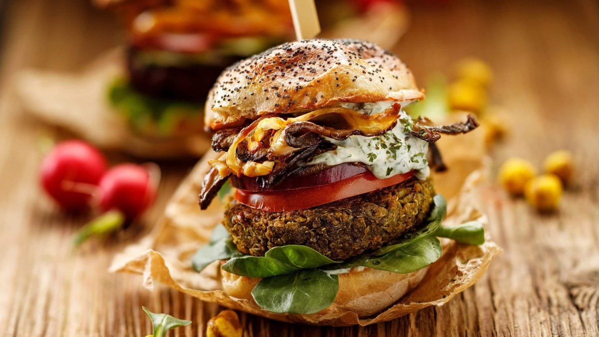 Erbsen-Burger in einem Sesambrötchen mit Tomate, Salat, frischer Remoulade, Röstzwiebeln und veganem Bacon auf einer hölzernen, rustikalen Unterlage