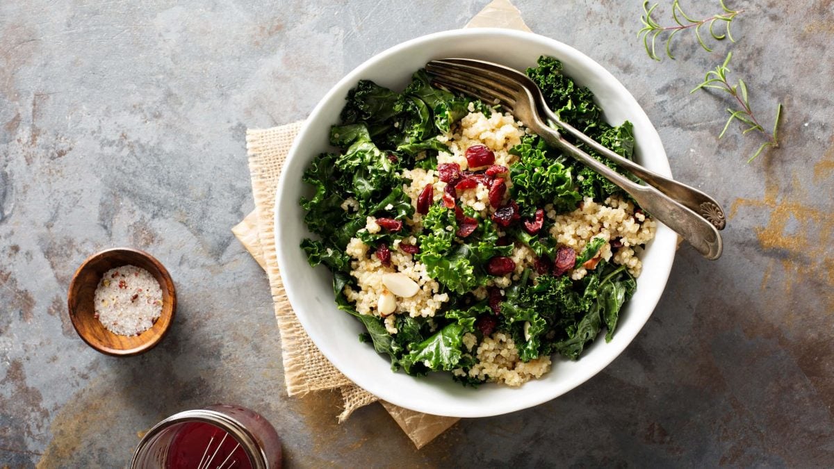 Eine weiße Schüssel Grünkohlsalat mit Quinoa und Cranberrys, darin eine Gabel und ein Löffel, in der Draufsicht. Daneben ein Schälchen Salz und ein Glas Dressing.