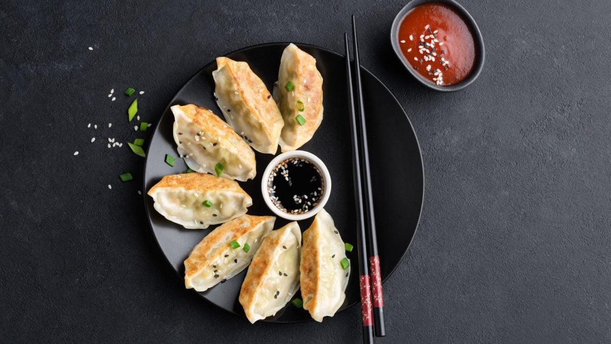 Gyoza mit Hähnchenfüllung mit schwarzen Essstäbchen auf schwarzem Teller und schwarzem Grund. In der Tellermitte eine weiße Schale mit dunklaer Sauce. Rechts oben schwarze Schale mit roter Sauce. Aufnahme aus der Vogelperspektive.