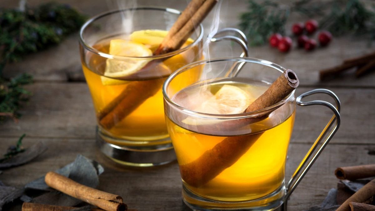 Zwei Hot Toddys mit Zitronenscheibe und Zimtstange in Glastasse auf Holztisch. Zimtstangen und Äste als Dekoration über dem Tisch verteilt. Aufnahme aus der Frontale.