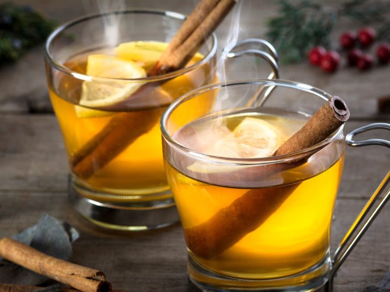 Zwei Hot Toddys mit Zitronenscheibe und Zimtstange in Glastasse auf Holztisch. Zimtstangen und Äste als Dekoration über dem Tisch verteilt. Aufnahme aus der Frontale.