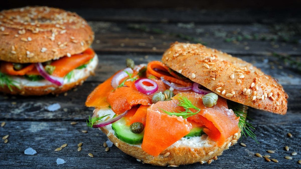 Veganer Karotten-Lachs mit Räuchersalz, Kapern und Öl auf einem Bagel mit Frischkäse, Gurke, Zwiebeln auf einem schwarzen Holzbrett
