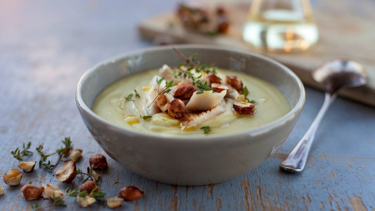Eine graue Schüssel Kartoffelsuppe mit Räucherfisch und Haselnüssen auf blauem Holz, daneben liegt ein Löffel.