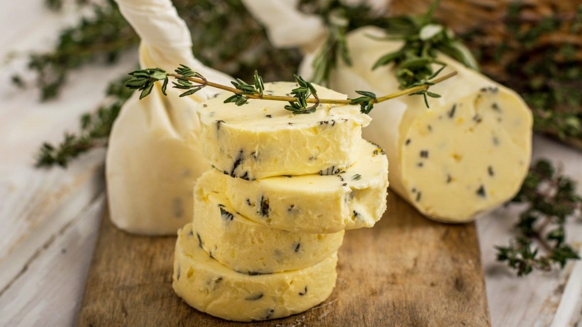 Auf einem Holzbrett liegt eine Kräuterbutter mit Limette. Die Butter ist in Pergamentpaier gerollt und mit Tyhmianzweigen dekoriert.