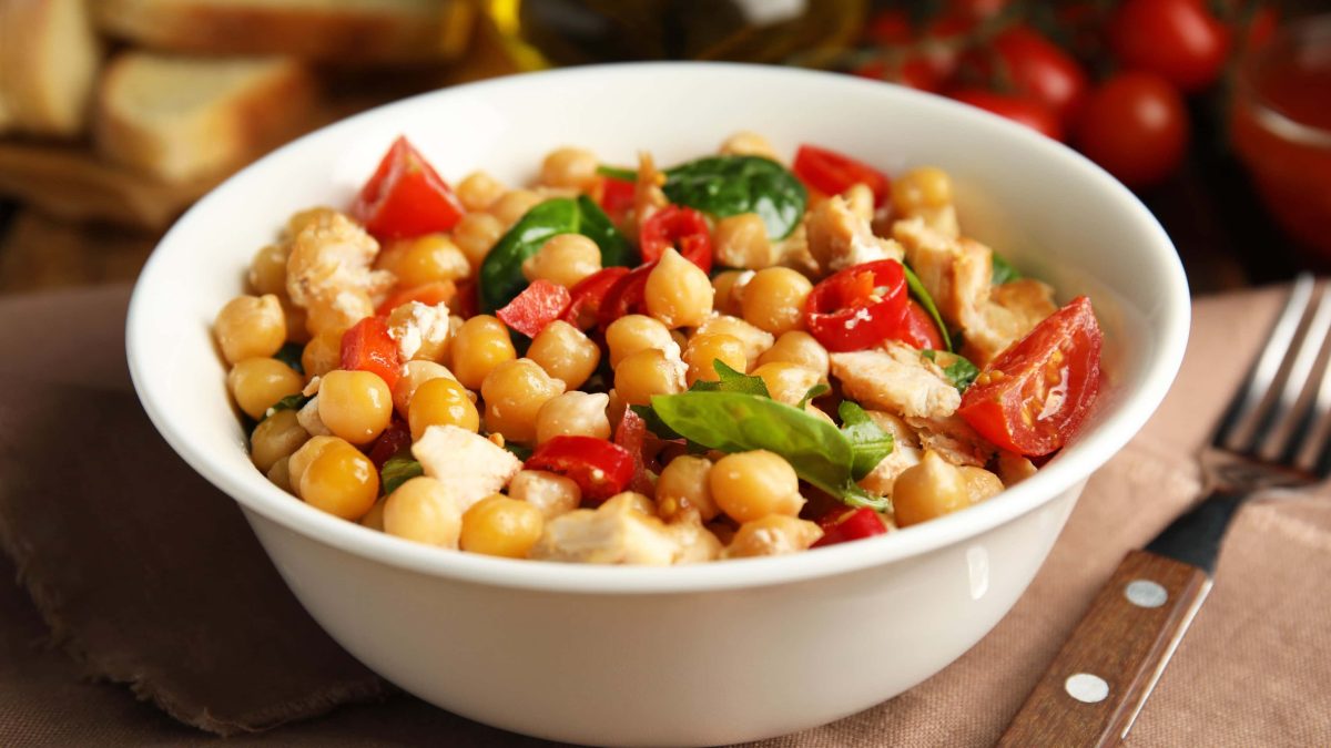 Kichererbsen, Halloumi, Spinat und Tomaten in weißer Schüssel. Daneben auf Holzbrett Gabel mit Holzgriff. Am oberen Bildrand unklar zu erkennen Brot und Datteltomaten.