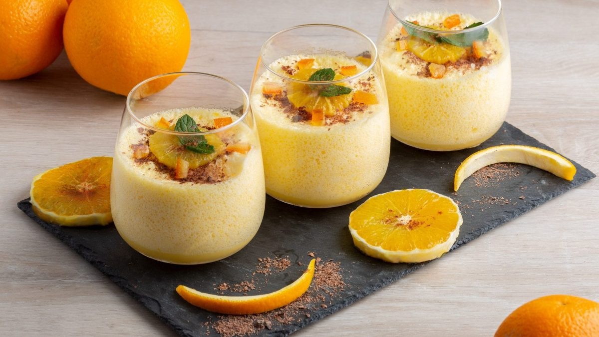 Auf einer Schiefertafel stehen drei Gläser mit Orangen-Mousse. Daneben liegen Orangen-Scheiben sowie drei Orangen.