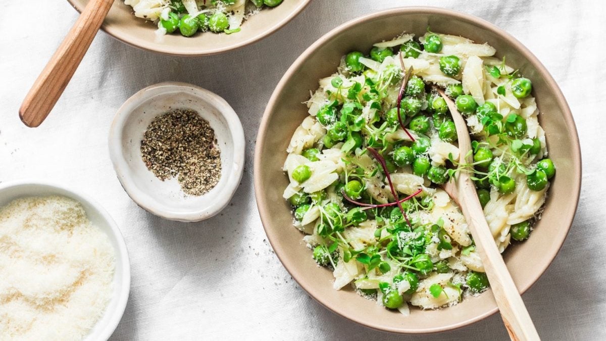 Ein tiefer Teller Orzonudeln mit Erbsen, grünem Pesto, Salat und geriebenem Käse, darin steckt ein Holzlöffel. Daneben zwei Schüsseln Pfeffer und Käse. Alles in der Draufsicht.