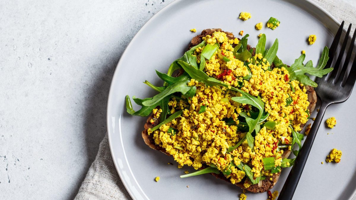 Würziges veganes Rühr-Ei, auch Rühr-Tofu genannt mit frischem Rucola und Brot auf einem weißen Teller und Gabel