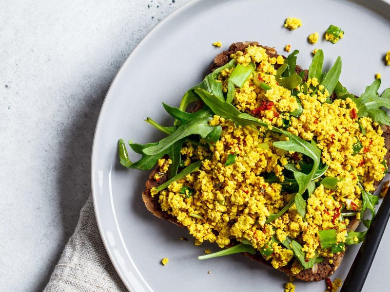 Würziges veganes Rühr-Ei, auch Rühr-Tofu genannt mit frischem Rucola und Brot auf einem weißen Teller und Gabel