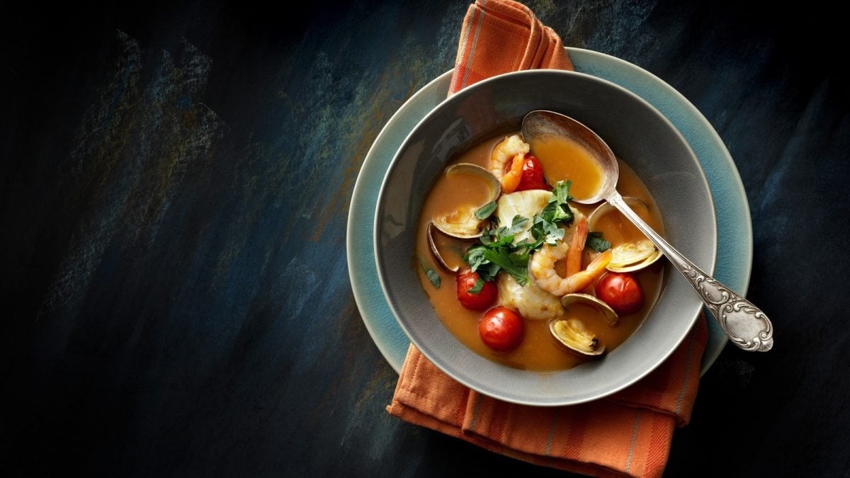 Eine Schüssel mit der Sopa de Pescados y Mariscos, der spanischen Fischsuppe, die mit Petersilie garniert ist, mit Löffel auf einem Unterteller und einer orangenen Stoff-Servierte vor einem dunklen Hintergrund, von oben.