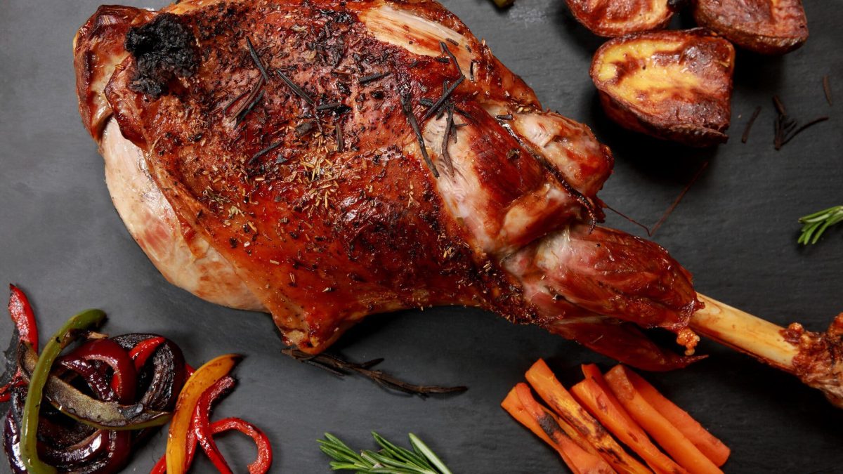Spanischer Lammbraten geschmortes Lamm mit Knochen auf grauer Platte. Links gebratene bunte Paprikastreifen, rechts gebratene Möhrenstifte. Gebacken, halbierte Kartoffeln oben rechts. Aufnahme aus der Draufsicht.