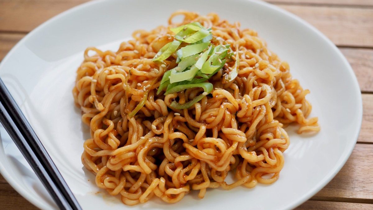 TikTok-Ramen mit Lauchzwiebel angerichtet auf einem weißen Teller. Der Teller steht auf einem Holztisch. Neben den Nudeln liegen zwei schwarze Esstäbchen.
