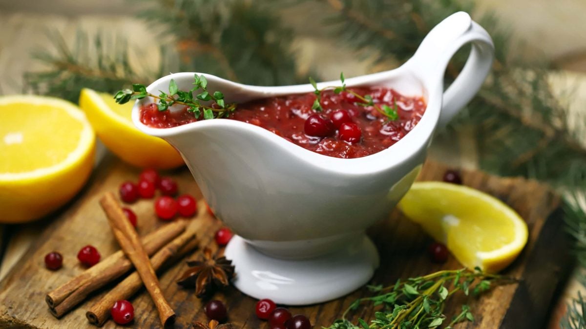 Eine Sauciere mit veganer Bratensauce und Cranberrys steht auf einem Holzbrett, drumherum liegen Orangenscheiben und Gewürze.