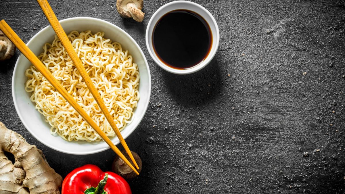 Ein Teller mit Ramennudeln und Essstäbchen, daneben steht eine Schale mit veganer Fischsauce. Alles in der Draufsicht auf dunklem Untergrund.