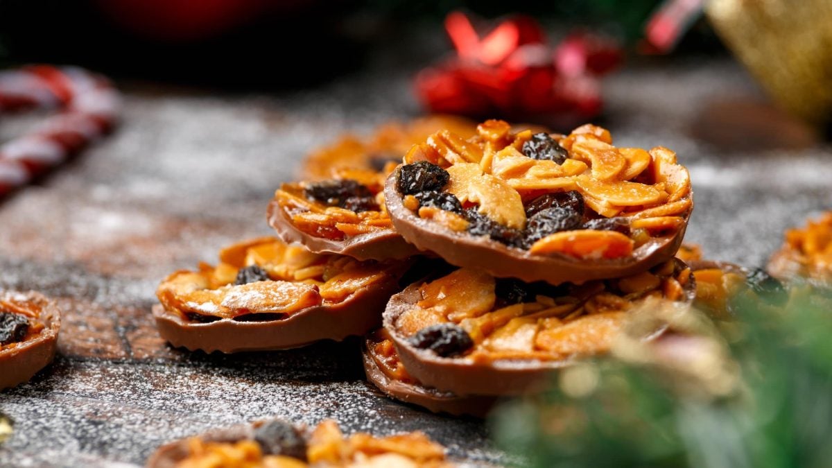 Auf einer mit Puderzucker bestäubten Fläsche liegen ein paar vegane Florentiner gestapelt. Im Hinter- und Vordergrund erkennt man unscharf etwas Weihnachtsdeko.
