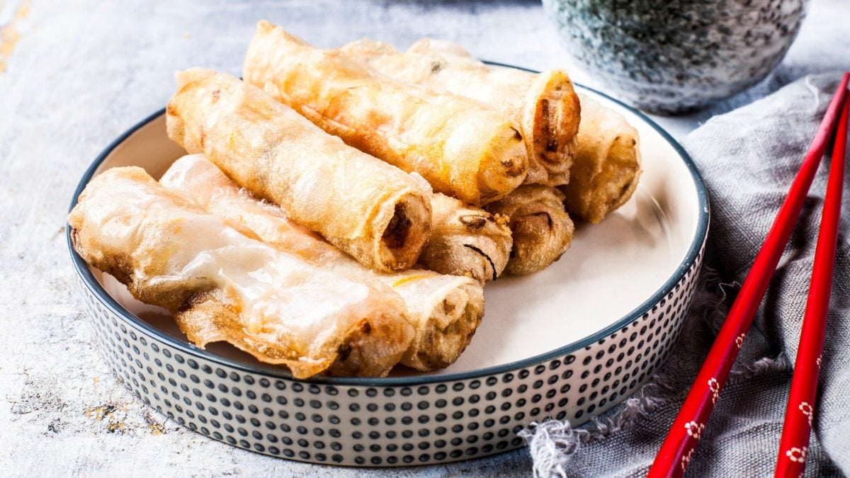In einer kleinen Schüssel liegen einige vegane Frühlingsrollen, daneben rote Essstäbchen.