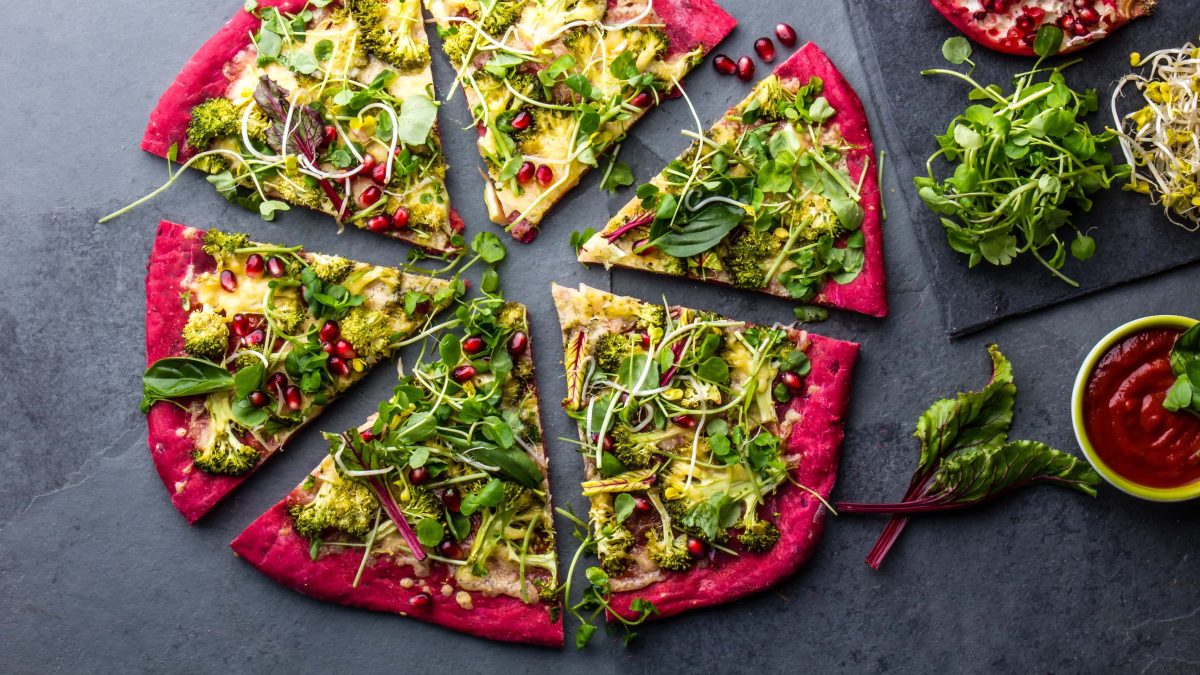 Eine geschnittene Rotkohl-Pizza mit üppigem Belag auf dunklem Untergrund in der Draufsicht, daneben liegen verschiedene Zutaten.