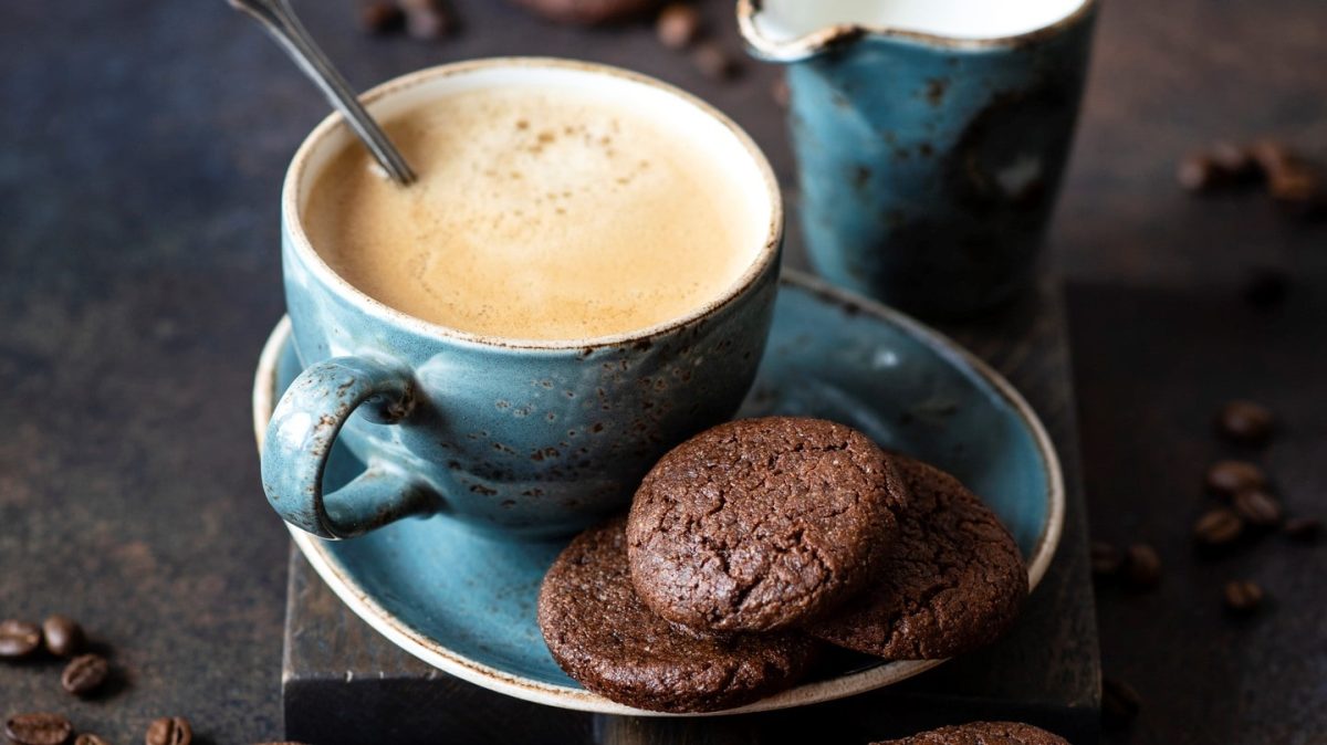 Eine gefüllte Kaffeetasse auf einer Untertasse, auf der einige vegane Schoko-Espresso-Plätzchen liegen. Drumherum verstreut sieht man Kaffeebohnen.