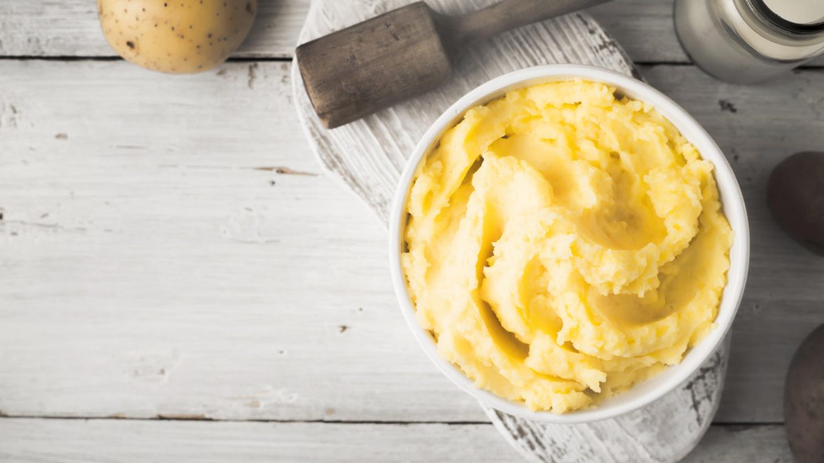 Eine Schale mit veganem Kartoffelpüree in der Draufsicht, sie steht auf einem weißen Holzbrett, daneben liegen ein Stampfer und eine Kartoffel.