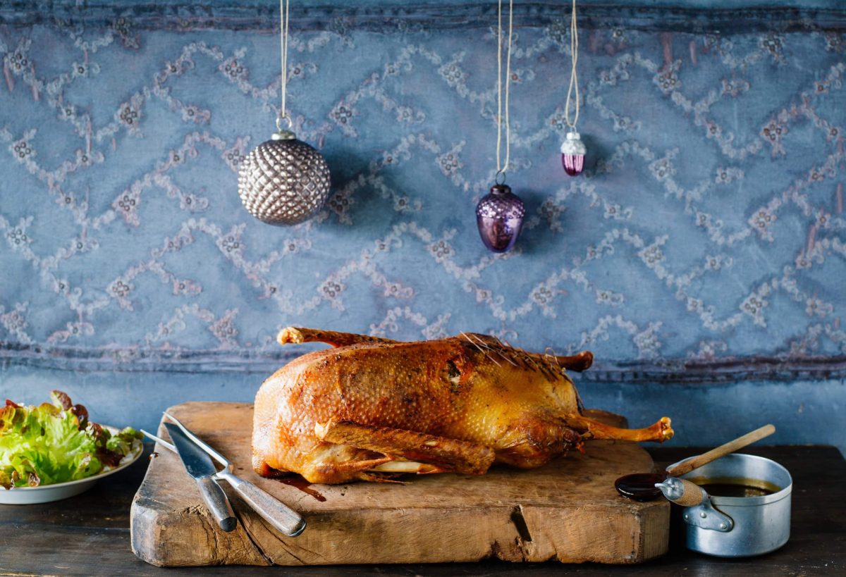 Die Weihnachtsgans liegt in der Küche auf einem dicken Holzbrett.. Im Hintergrund hängen Christbaumkugeln.