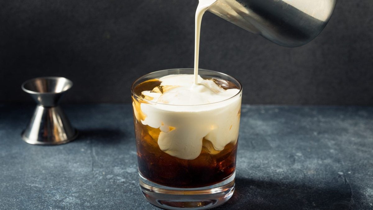 White Russian aus "The Big Lebowski" in der Entstehung im Tumbler auf Granittisch aus der Frontalansicht. Metallkännchen schüttet Sahne in das Glas. Links im Hintergrund Barmaß.