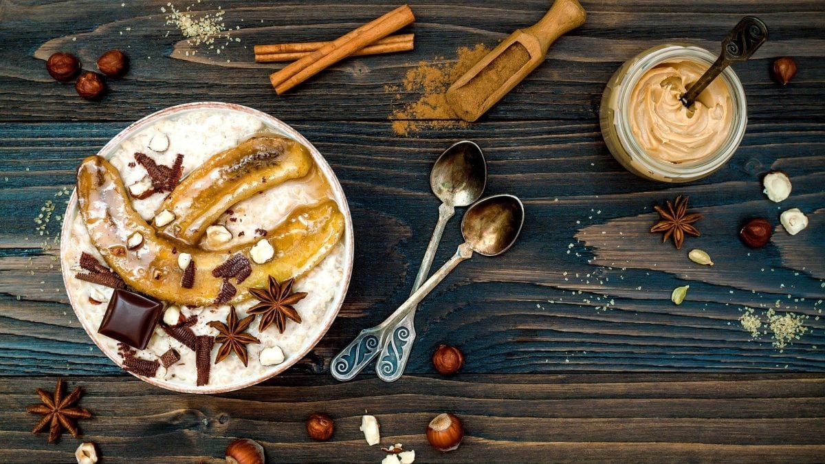 Ein winterlicher Porridge mit gebackenen Bananen steht auf einem dunklen Untergrund. Der Porridge ist angerichtet mit Schokolade, Zimt und Sternanis. Auf dem Tisch verteilt liegen Nüssem Löffel und Gewürze.
