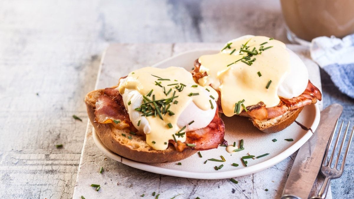 Eggs Benedict mit Speck auf Brot und Hollandaise, angerichtet auf einem weißen Teller.