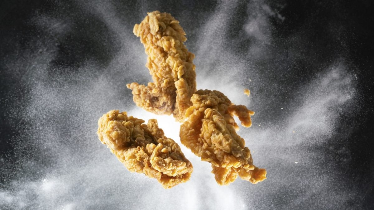 Drei panierte Chicken Wings, die in die Luft geworfen wurden. Im Hintergrund wirbelt weißes Mehl auf schwarzer Fläche.