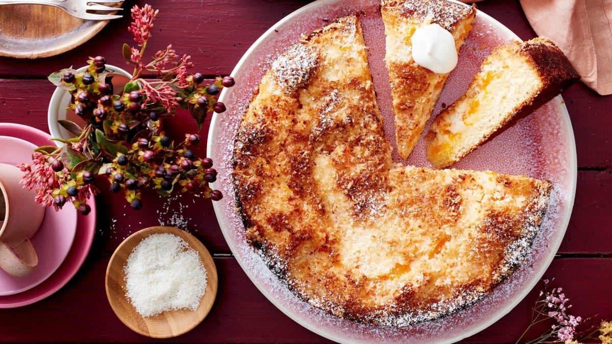 Auf einem Teller liegt ein fluffiger Mandarinen-Kokos-Kuchen. Er ist mit Puderzucker bestreut und mit einem Klecks Schlagsahne verfeinert.