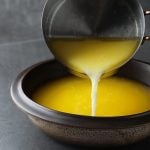 Vor einem dunklen Hintergrund ist eine SChale und ein Topf zu sehen. Aus dem Topf wird eine gelge Flüssigkeit in die Schale geschüttet. Bei der Flüssigkeit handelt es ich um geklärte Butter.