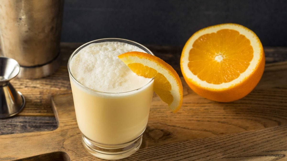Orange Whip aus "Blues Brothers" in Longdrinkglas mit Orangenschale auf Holztisch. Daneben rechte eine halbe Ornae mit der Schnittseite zum Betrachter. Link Cocktailzubehör. Aufnahme aus der Frontalen.