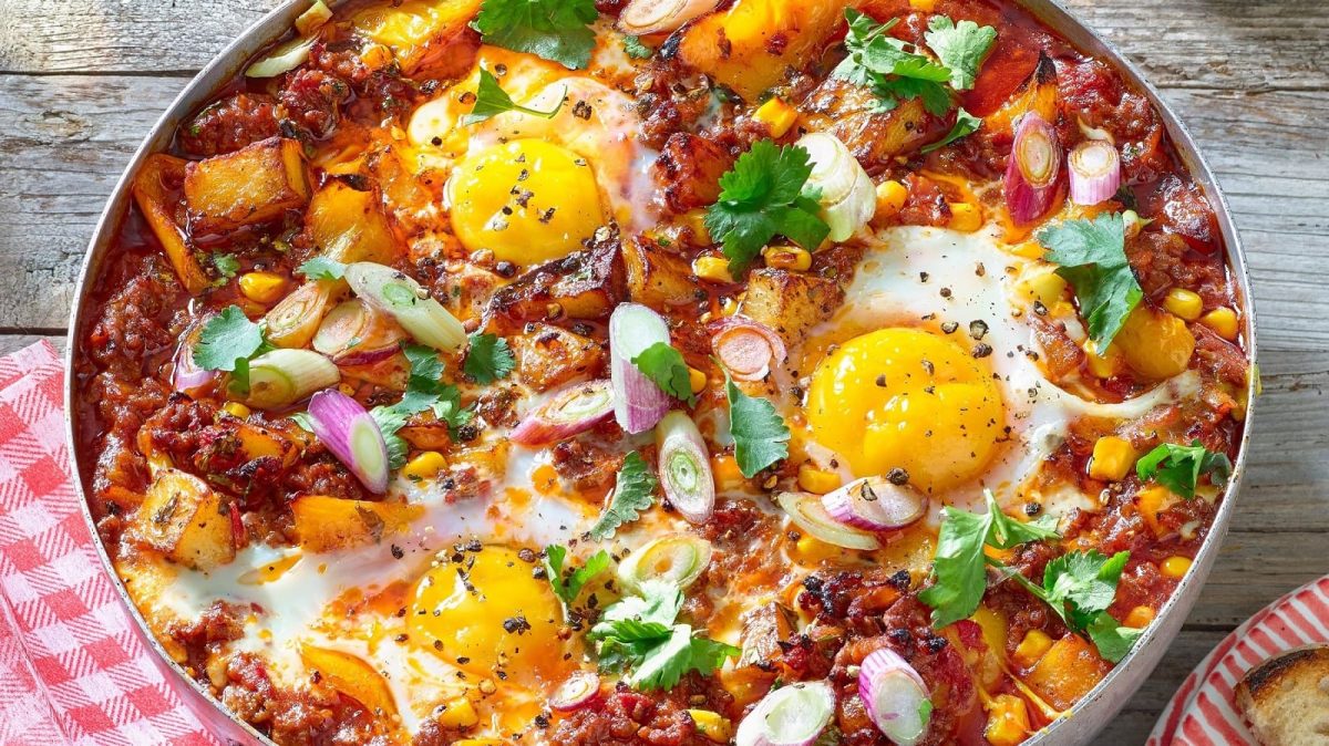 Eine große Pfanne mit dem Bolognese-Shakshuka, garniert mit Korianderblättern und Frühlingszwiebeln, bestreut mit frisch gemahlenem Pfeffer neben einem rot-weiß karierten Geschirrtuch und einem Teller mit einem Stück Brot, vor einem Holzhintergrund von oben.