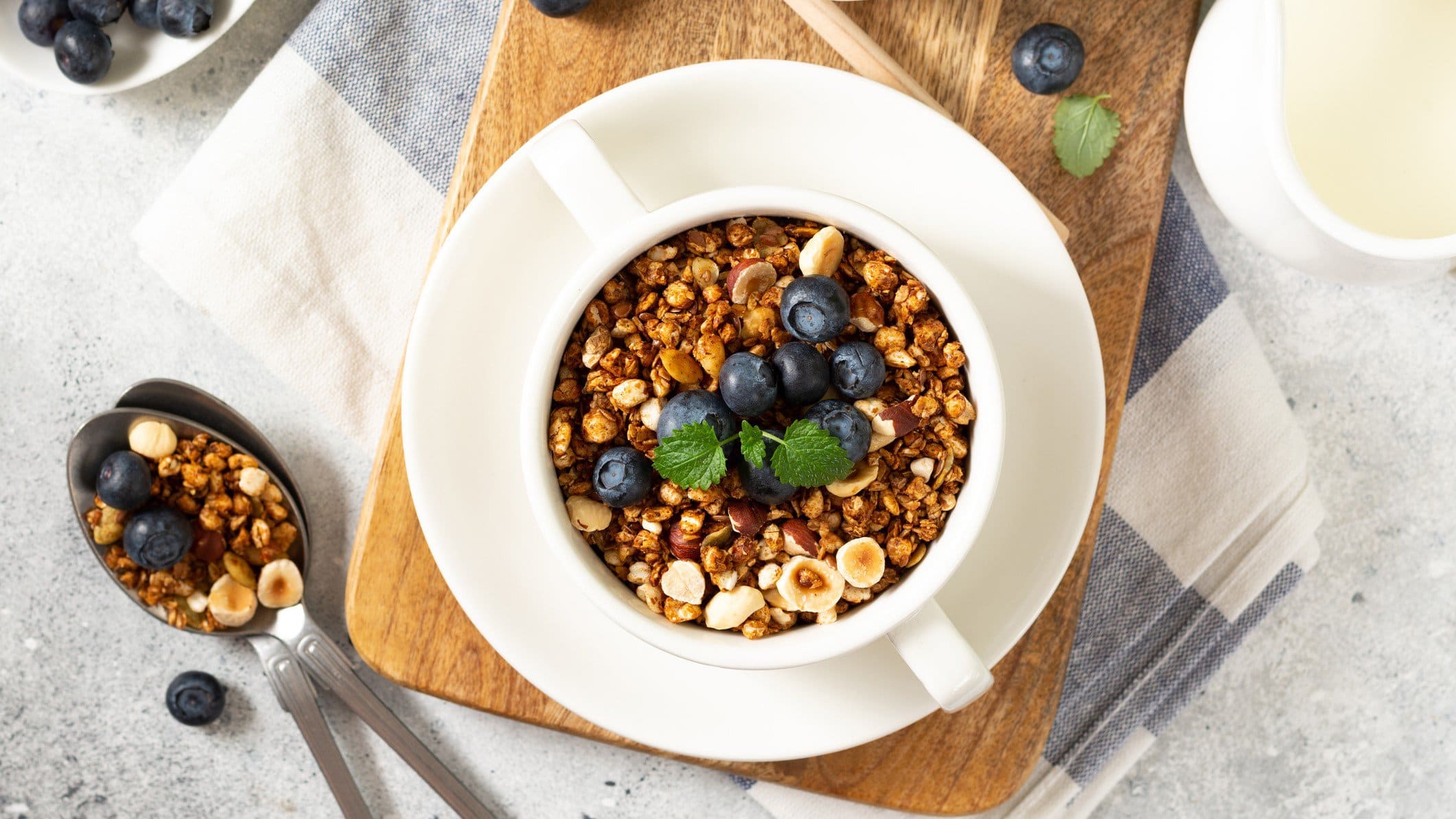 Eine Schale Buchweizenmüsli mit Haselnüssen und Blaubeeren in der Draufsicht, daneben ein Löffel Müsli und ein Kännchen Milch.
