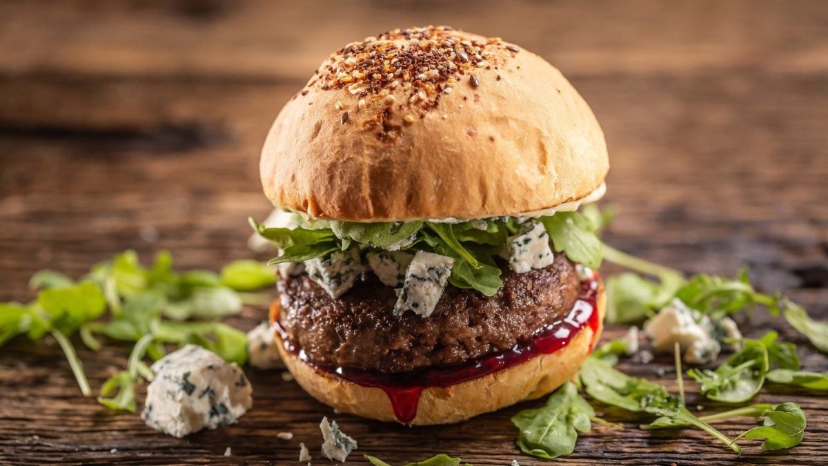 Ein Burger mit Blauschimmelkäse und Preiselbeersauce auf einem Untergrund aus dunklem Holz. Um den Burger herum liegen noch Rucola und Blauschimmelkäse.
