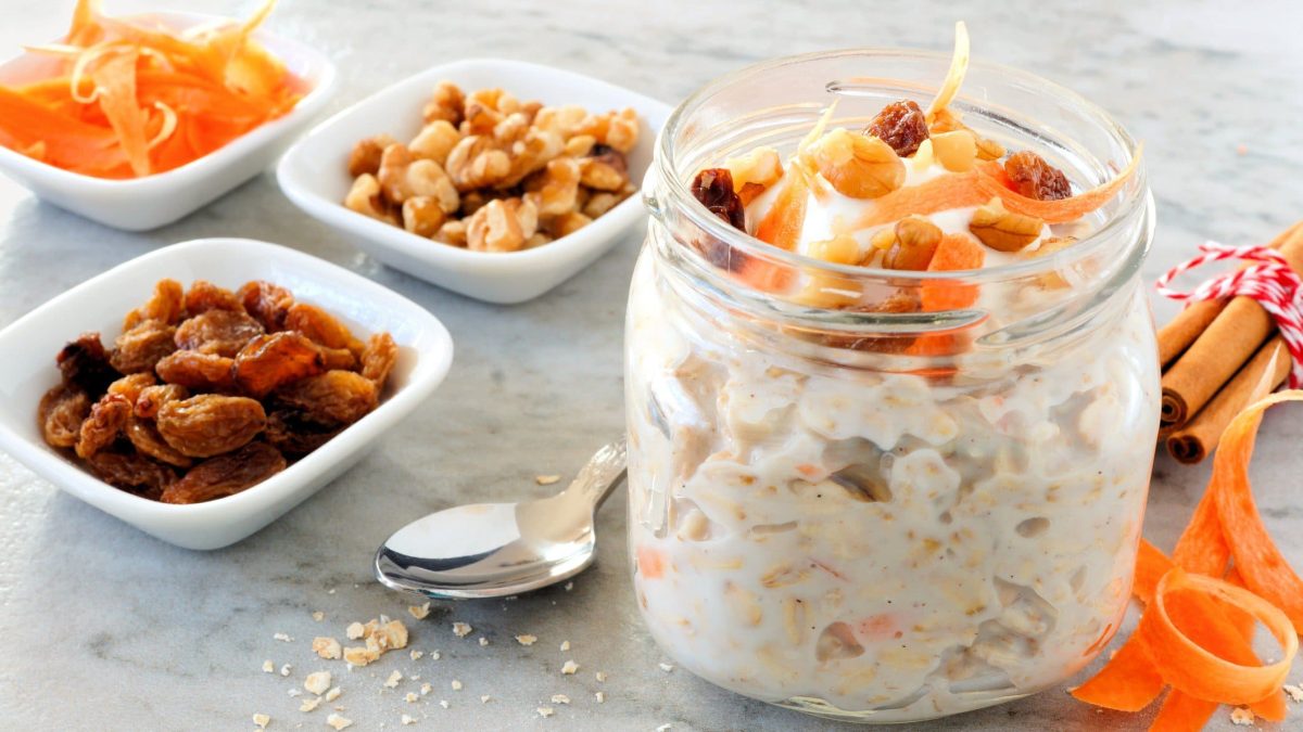 EIn Glas Carrot Cake Overnight Oats, daneben stehen Schälchen mit Zutaten wie Nüssen oder Rosinen, außerdem geraspelte Möhren.