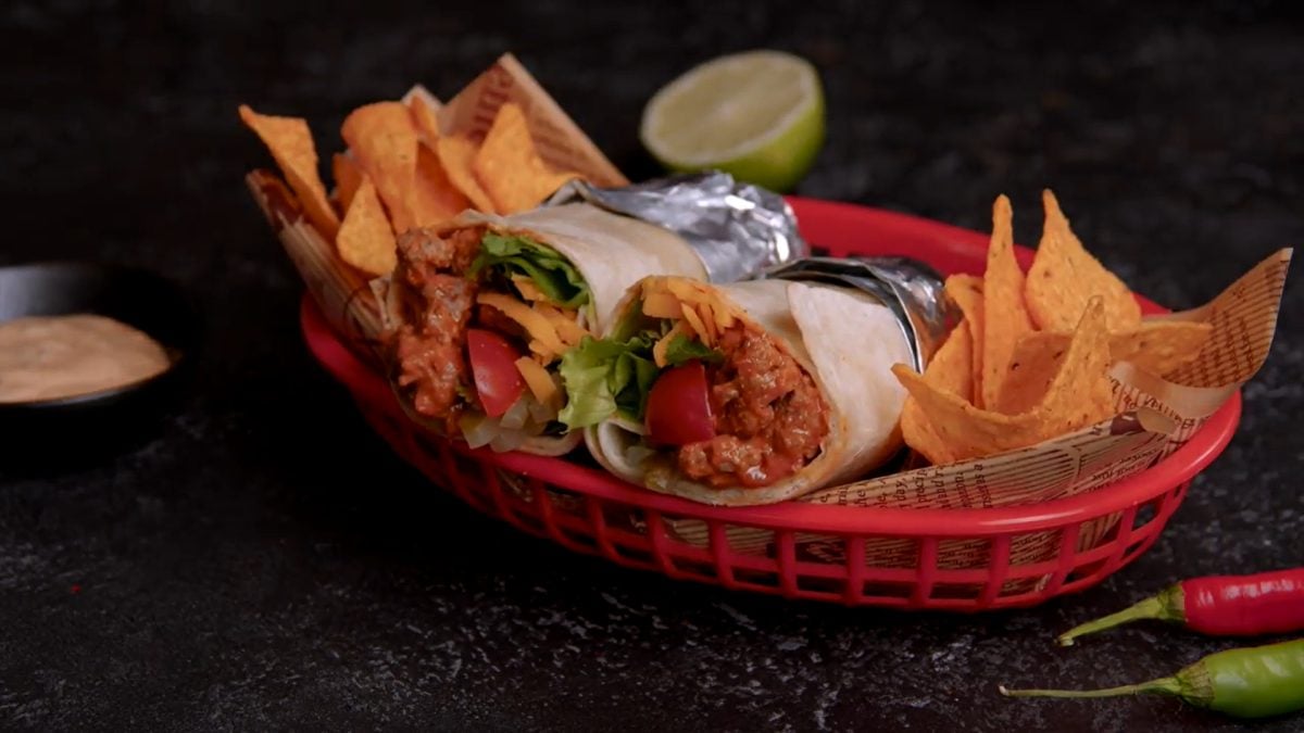 Cheeseburger-Burrito in aufgeschnitten in Alufolie gewickelt mit Nachos in rotem Brotkorb. Schnittseit zum Betrachter. Rechts eine grüne und eine rote Chili. Halbe Limette oben und Dip links im Bild. Frontalaufnahme.