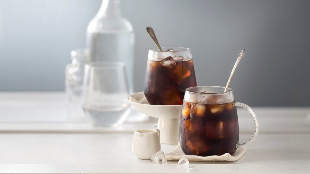 Zwei Gläser Coffbucha mit Eis in einem hellen Setting, im Hintergrund unscharf eine Flasche Wasser und Gläser.