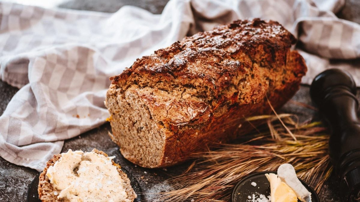 Ein angeschnittenes Dinkelbrot mit Nüssen, daneben liegen Getreideähren, ein Geschirrtuch und Butter.