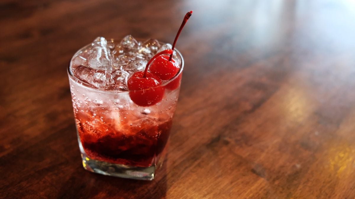 El Diablo Longdrink Cocktail im kleinen Longdrink-Glas mit Cocktailkirschen auf Holztisch. Frontalaufnahme.
