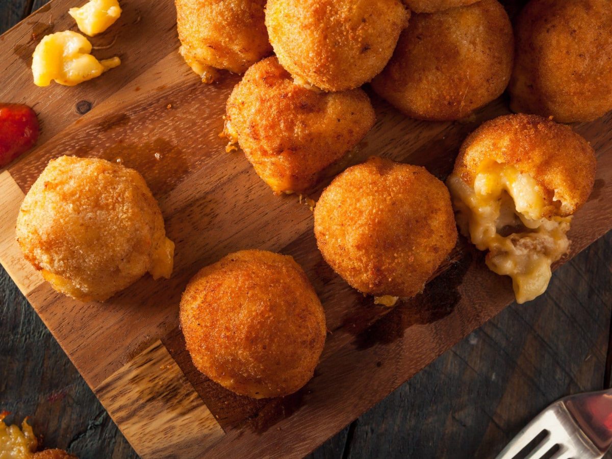 Fried Mac'n Cheese Balls frittierte Maccaroni mit Käse auf Holzbrett und dunklem Tisch. Ketchup und einzelne Maccaroni auf dem Brett. Rechts unten Messer und Gabel. Aufnahme aus der Vogelperspektive.