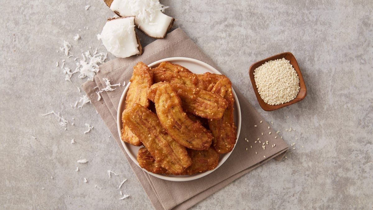 Ein Teller frittierter Bananen in der Draufsicht, daneben sind Kokosnussstücke und eine Schale mit weißem Sesam zu sehen.