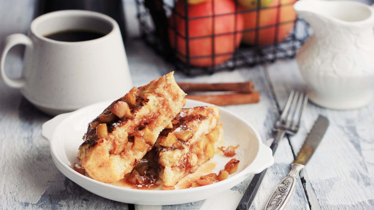 Ein Teller gefüllte French Toasts mit Apfelkompott, daneben Besteck, eine Tasse Kaffee und ein Pott Milch.