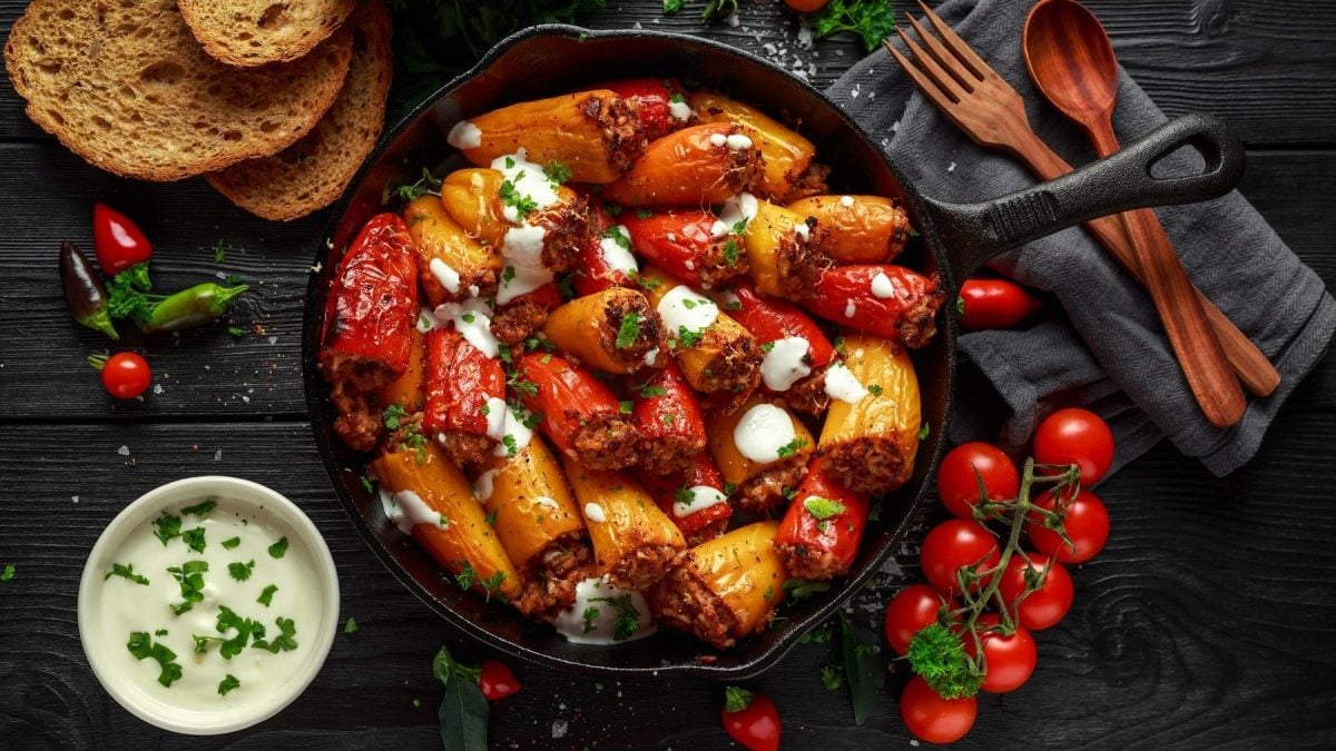 Aus der Vogelperspektive: In einer PFanne sind rote und gelbe Mini-Paprika, die mit Lamm und Feta gefüllt sind. Drumherum liegen Tomaten, ein paar Scheiben Brot, eine SChale mit Joghurt sowie ein Besteck.
