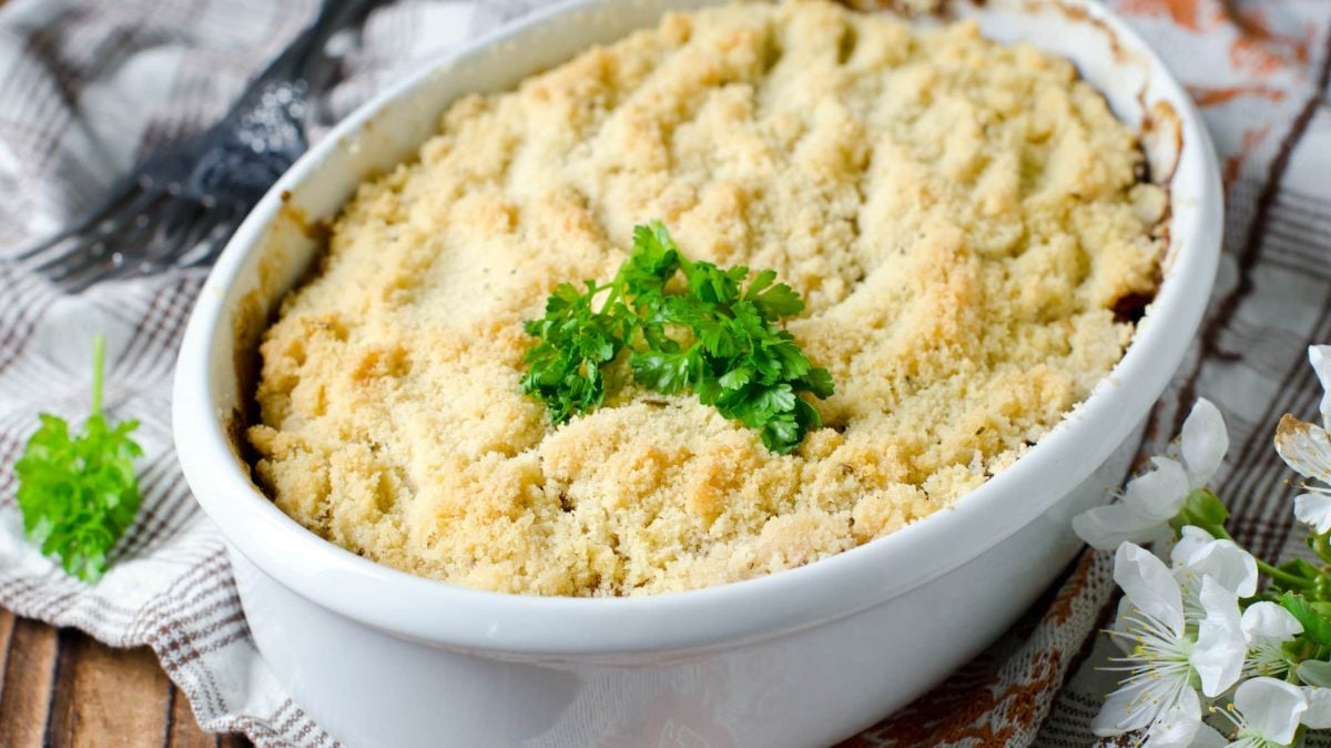 Eine weiße Auflaufform Gemüse-Crumble mit frischen Kräutern dekoriert.