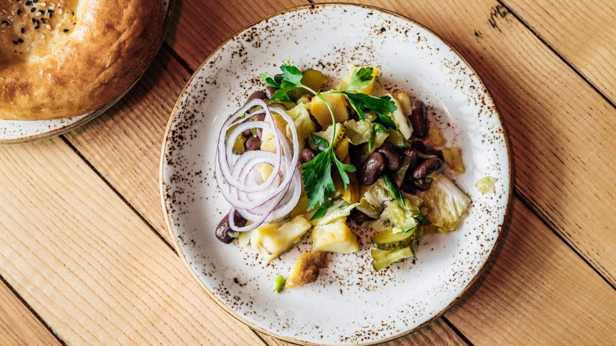 Ein würziger, saftiger georgischer Kartoffelsalat aus Kartoffeln, saurer Gurke, Kidneybohnen, Kohl, Zwiebeln und Petersilie auf einem hellen Tonteller auf hölzernem Untergrund, serviert mit frischem Fladenbrot