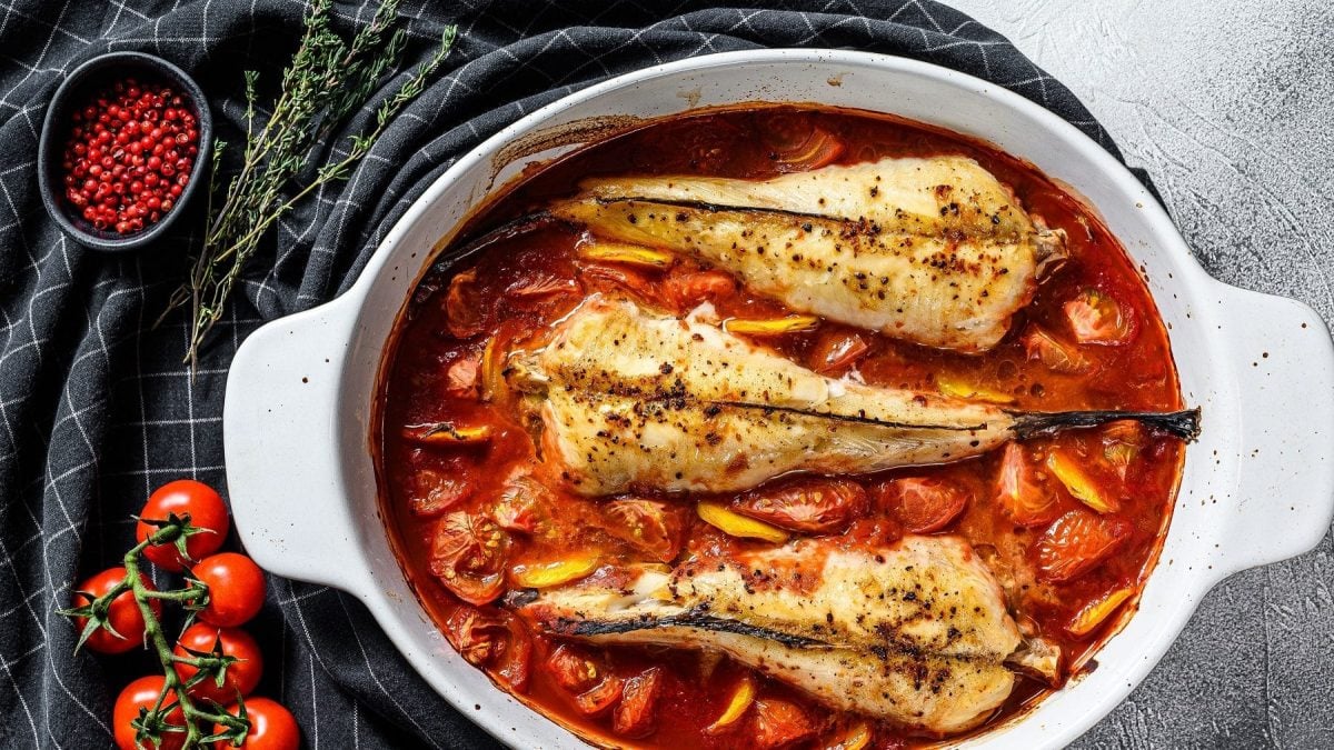 Au der Vogelpersektive: In einem großen Schmortopf liegen drei Seeteufel am Stück, aber ohne Kopf. Seeteufel sind Fische. Diese liegen in einem Bett von Tomaten und Paprika und Rosmarin.
