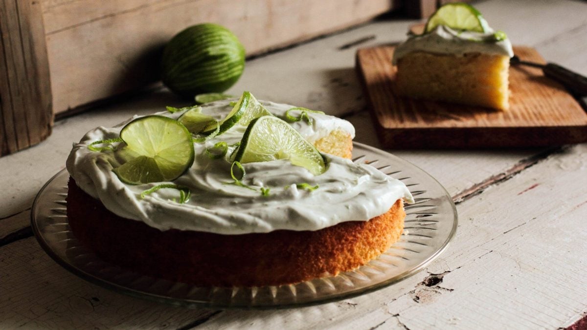 Ein angeschnittener Gin-Tonic-Kuchen auf einer runden Kuchenplatte, dahinter steht ein Stück vom Kuchen auf einem Holzbrett.