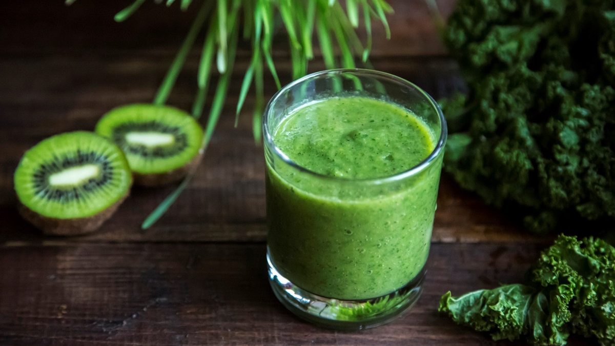 Ein Glas mit Grünkohl-Smoothie. Auf dem dunklen Untergrund liegen Kiwis und Grünkohl.