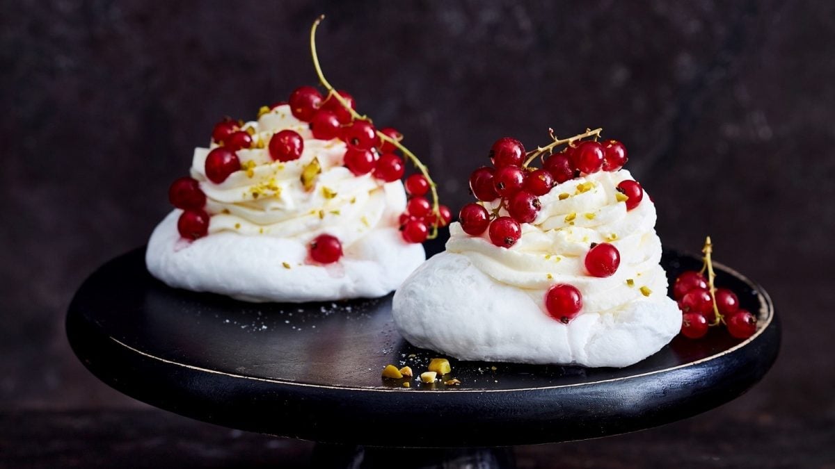 Eine schwarze Platte auf der zwei Johannisbeer-Baisertörtchen garniert mit Pistazien und frischen Johannisbeeren, zu sehen sind, vor einem dunklen Hintergrund.