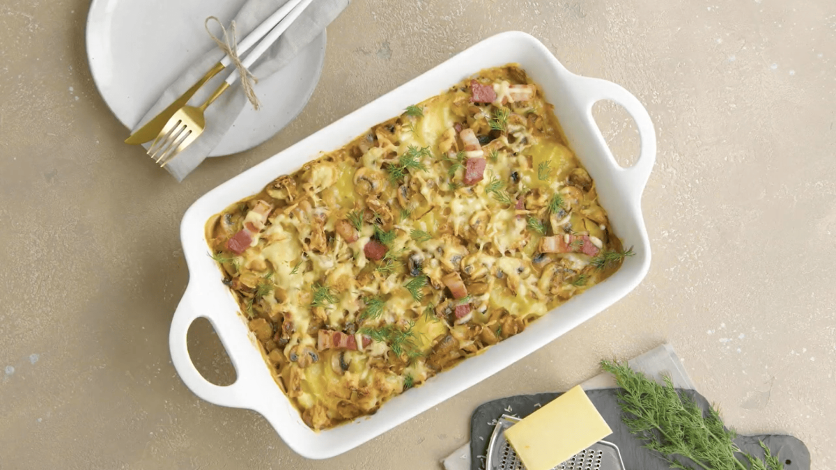 Eine weiße Auflaufform Kartoffelauflauf mit Speck und Pilzen in der Draufsicht, außerdem ein Teller mit Besteck sowie ein Stück Käse und eine Reibe.